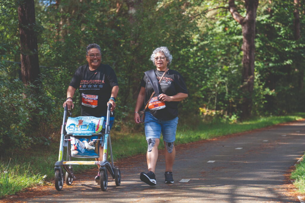 Twee wandelaars met startnummer lopen lachend in het bos waarvan één met een rollator en de ander met knietape